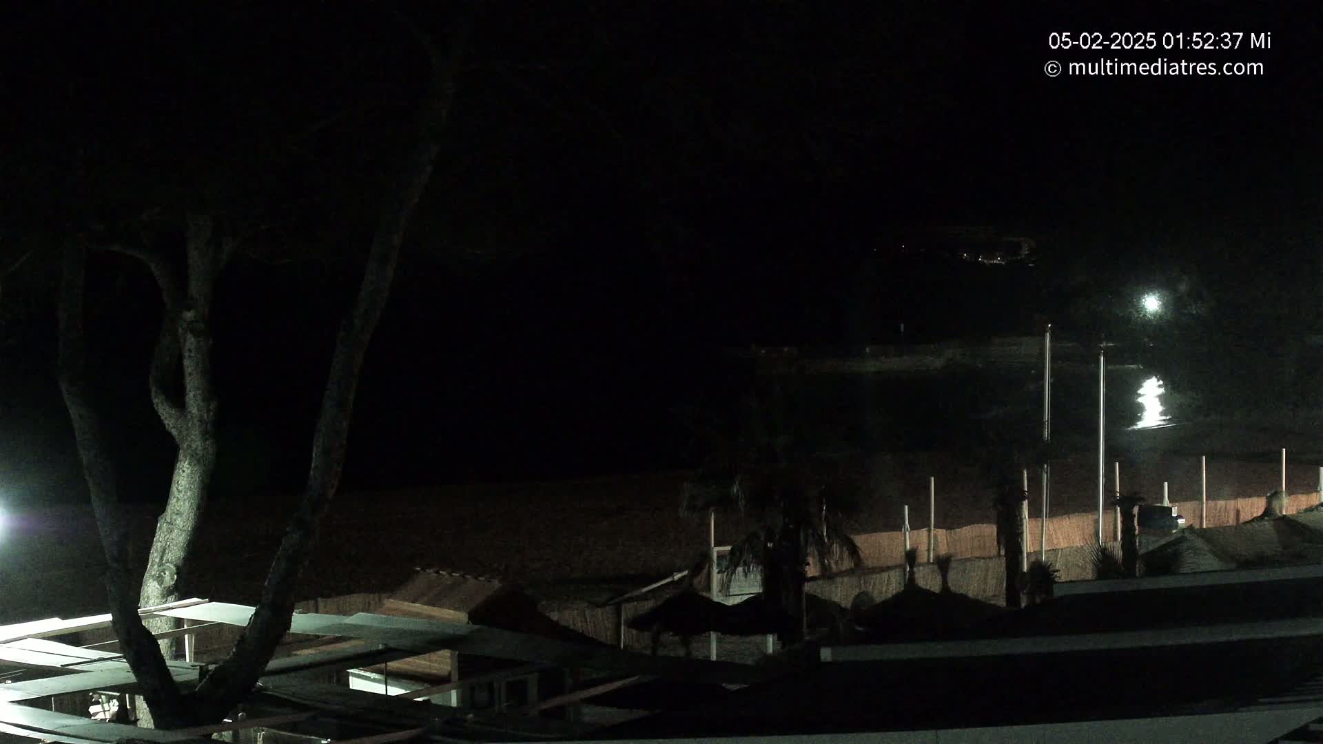 A dark, nighttime view of a beach with structures and a tree, under a mostly clear sky.