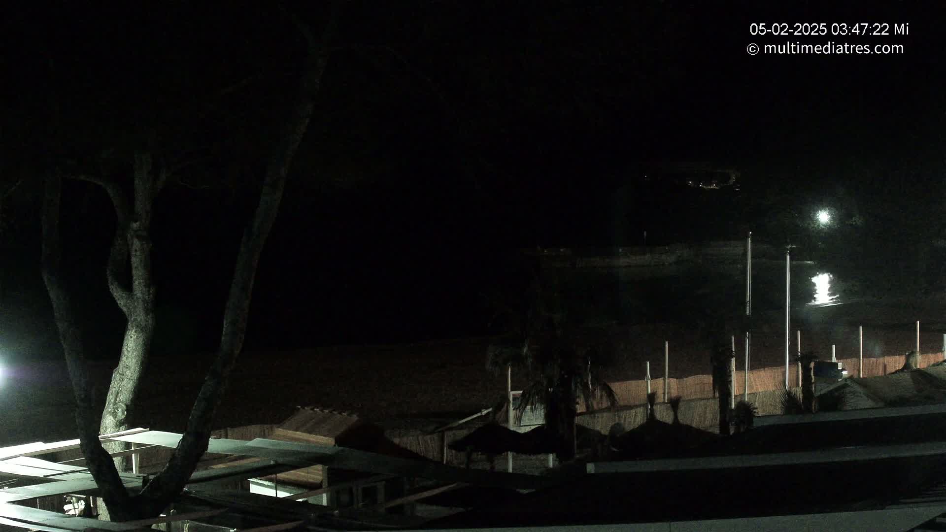 A nighttime view of a dark beach with several structures and palm trees, illuminated by artificial lights.
