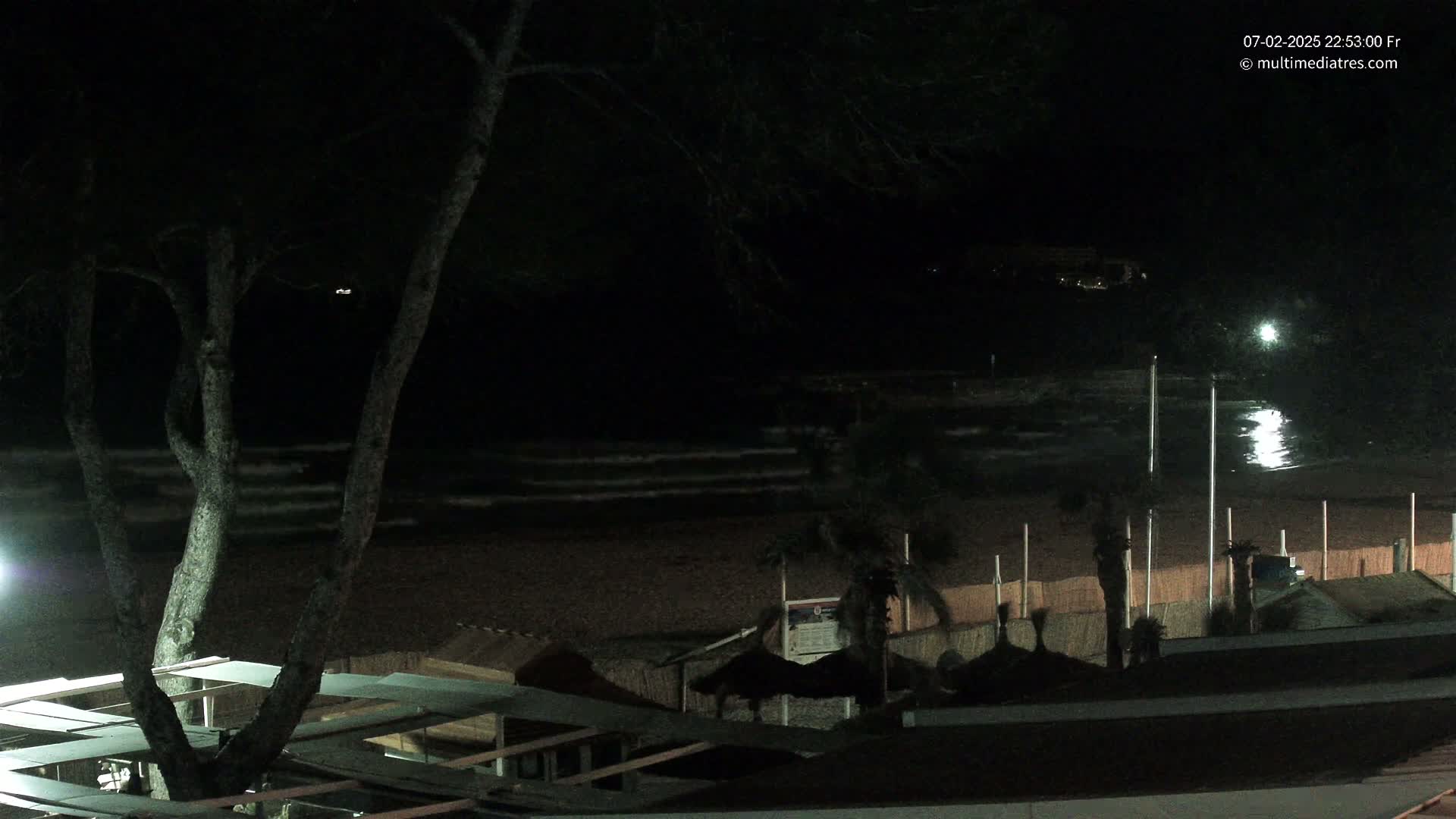 A dark nighttime beach scene shows gently lapping waves, a sandy shore, and beachside structures under a mostly clear sky.