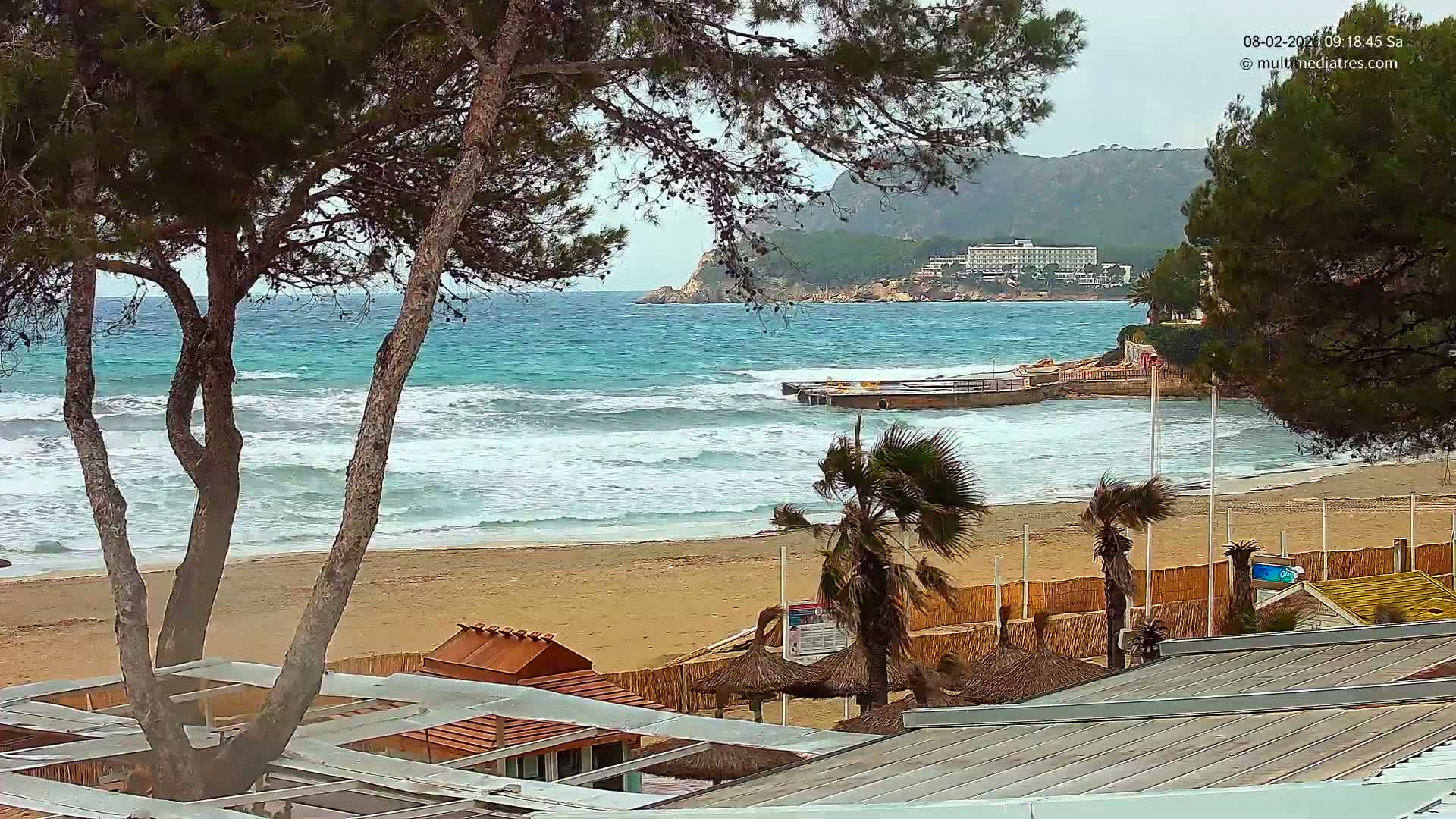 A sandy beach with moderate waves and some palm trees is seen from above, with a hotel visible in the distance on a cloudy day.