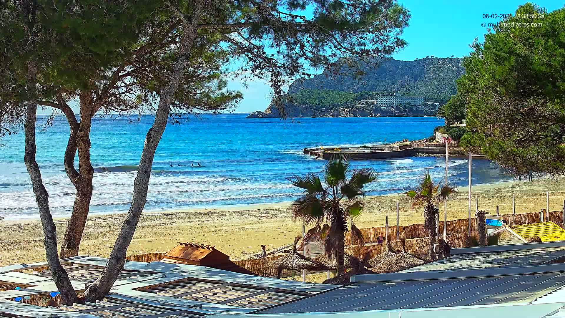 A sandy beach with calm waves and turquoise water is viewed from behind beachside structures and pine trees, under a sunny sky with a rocky cliff in the distance.