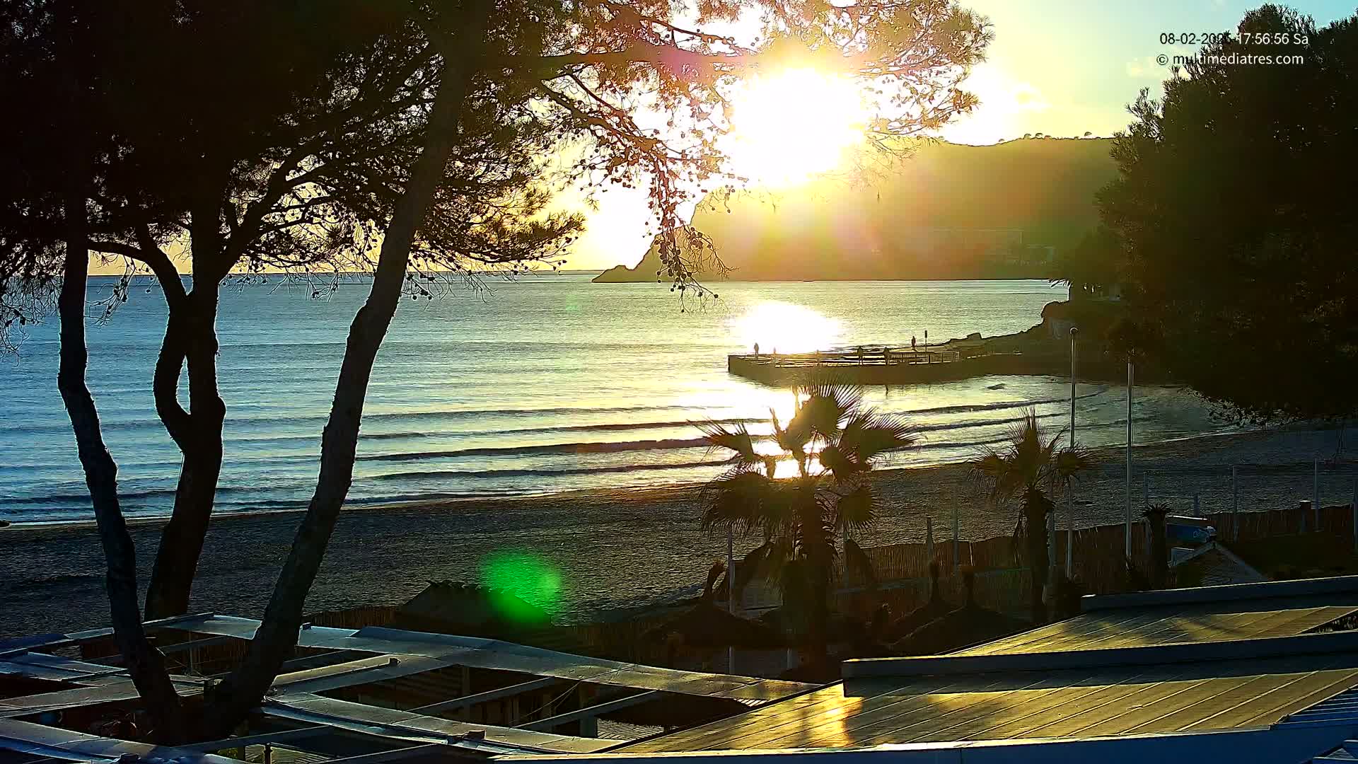 A sandy beach with gentle waves, palm trees, and beach structures is bathed in the golden light of a sunrise, with a hill visible in the background.