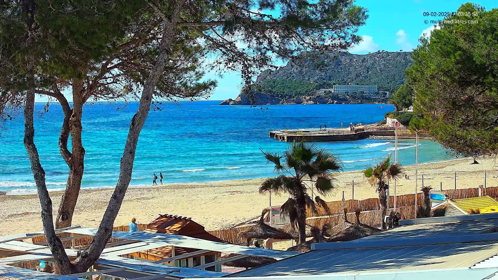 A sandy beach with turquoise water, palm trees, and a few people is seen from behind a row of wooden structures on a sunny day.