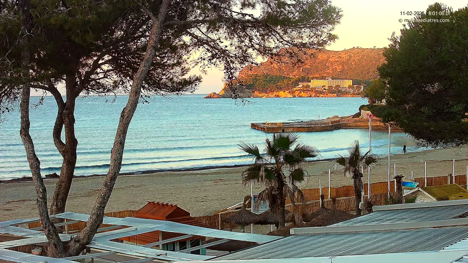 A sandy beach with calm waves, palm trees, and beach structures is seen from above at sunrise, with a distant view of a hill and buildings across a calm bay.
