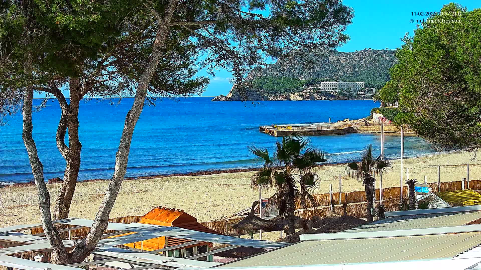 A sandy beach with turquoise water, palm trees, and beachside structures is visible under a clear blue sky.