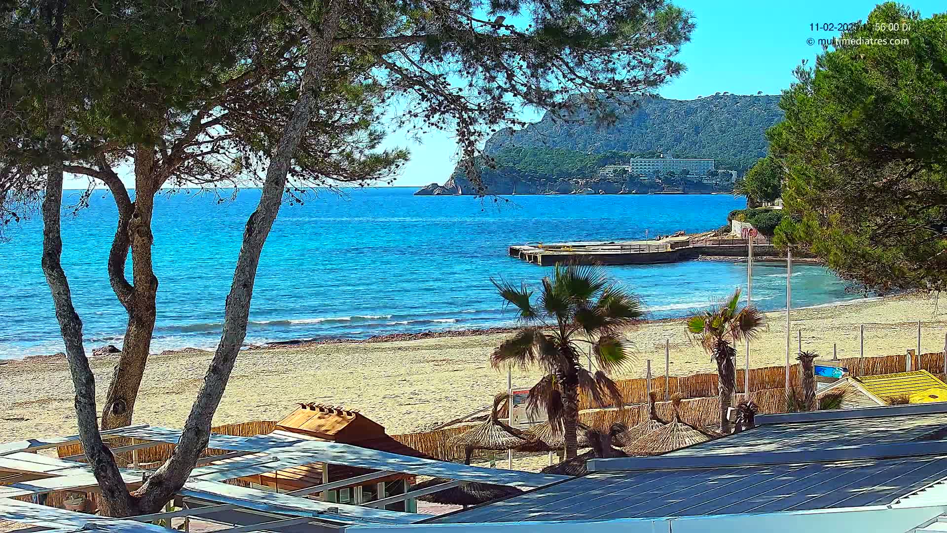 A sandy beach with turquoise water, palm trees, and beach umbrellas is seen from behind a row of buildings on a sunny day.