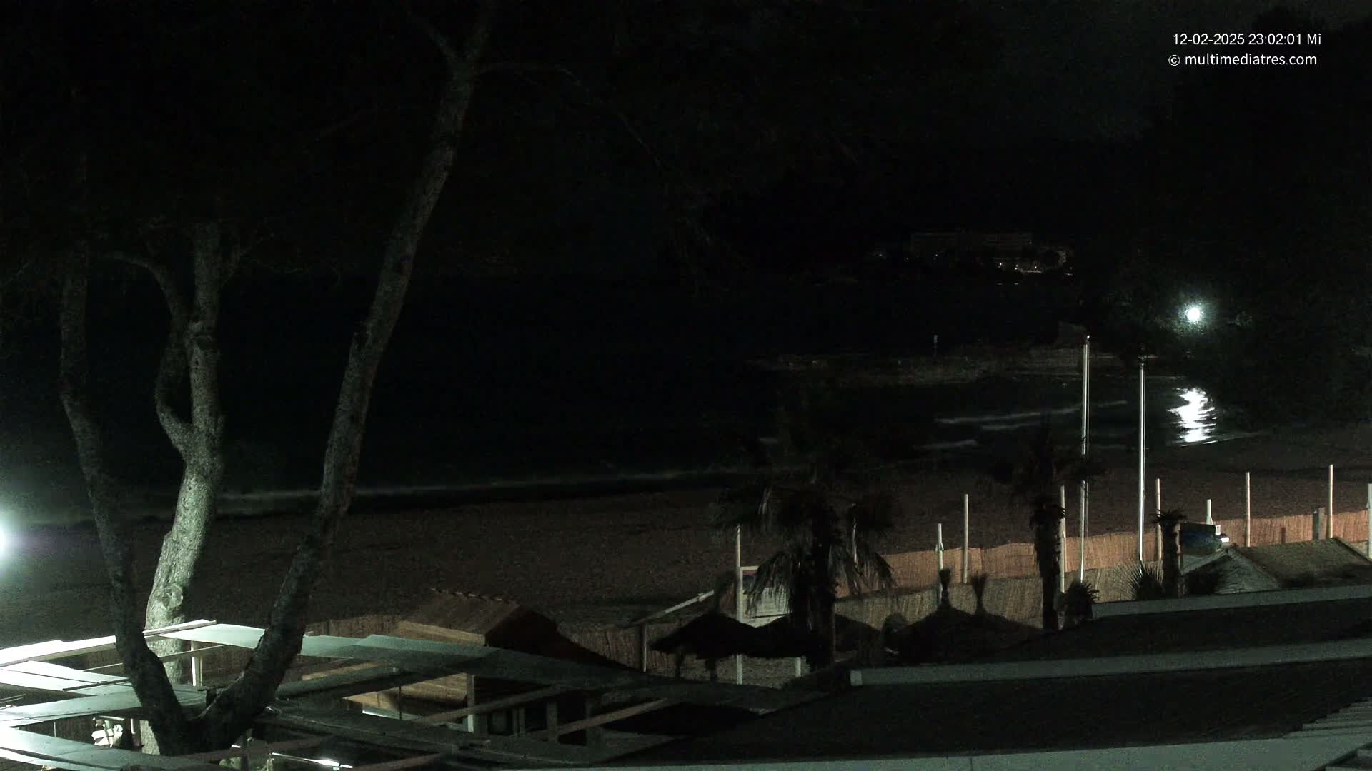 A nighttime view of a dark beach with gentle waves, palm trees, and several small structures under a mostly clear sky.