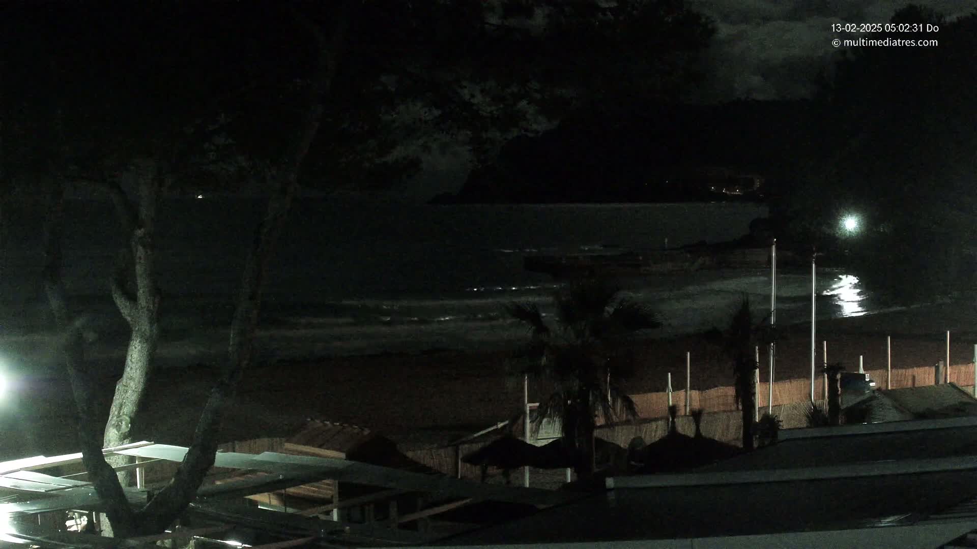 A nighttime view of a dark beach with gently lapping waves under a mostly cloudy sky, showing palm trees and beachfront structures.