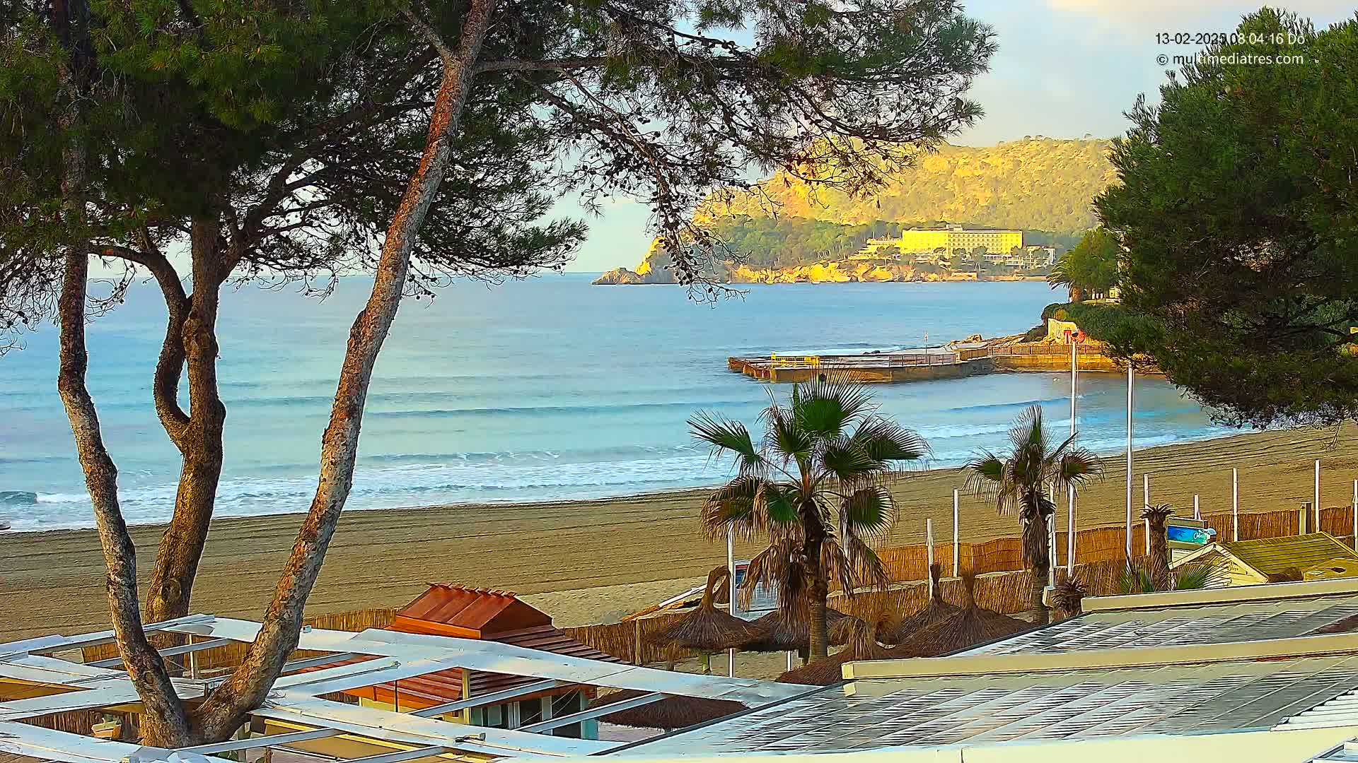 A sandy beach with gentle waves laps the shore under a calm, clear sky, with palm trees and beach structures in the foreground and a distant hillside visible beyond the water.