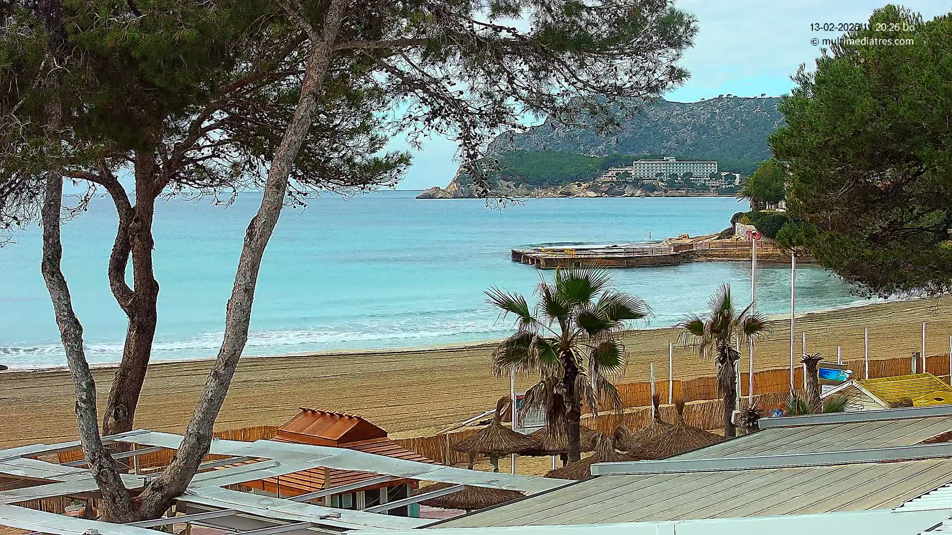 A sandy beach with calm, turquoise water is viewed from behind beachside structures and palm trees under a mostly cloudy sky.