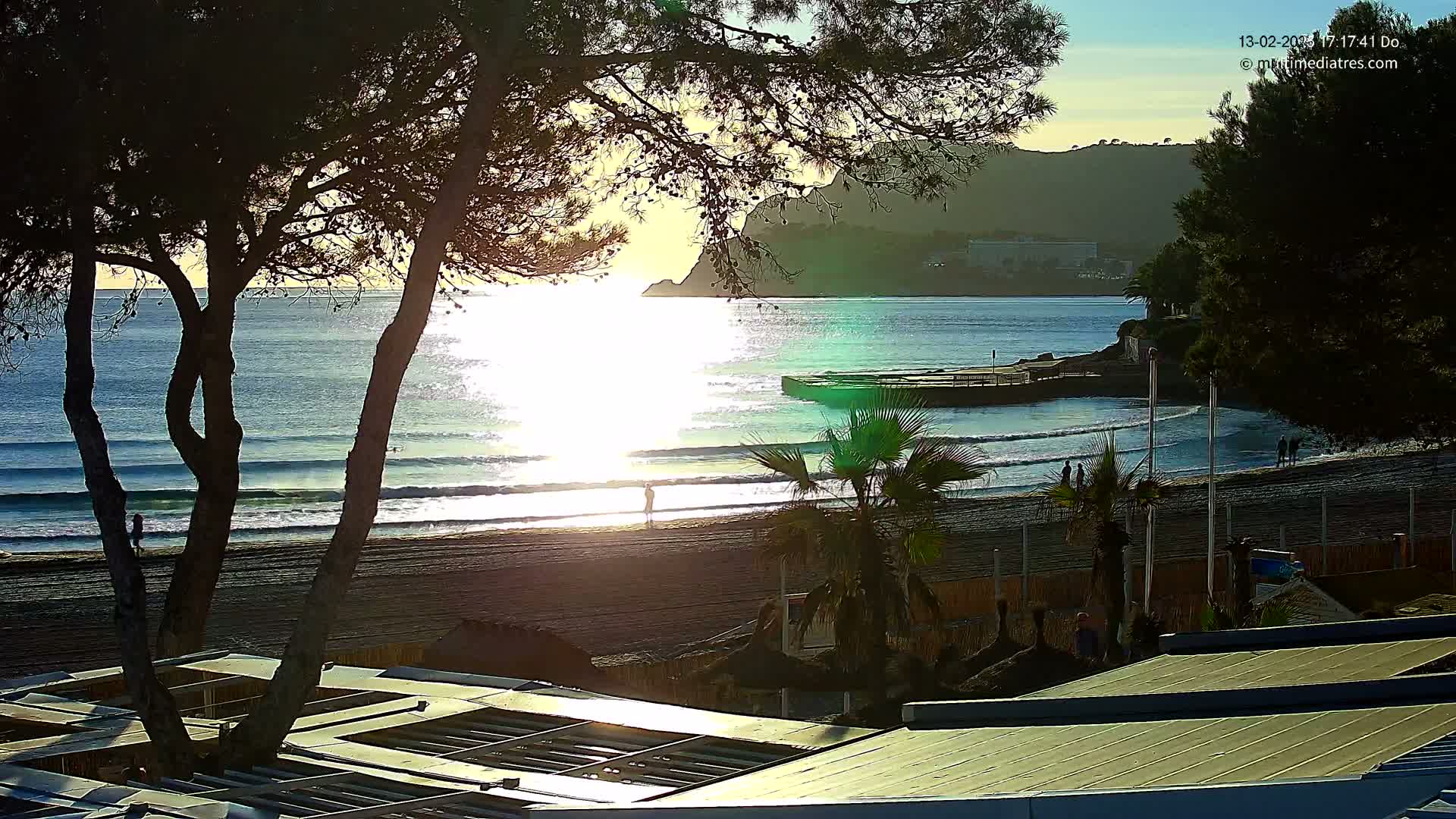 A sandy beach with gentle waves laps the shore under a clear, bright sky, as the sun shines on the water, seen through the branches of dark trees and partially obscured by a building’s roof.