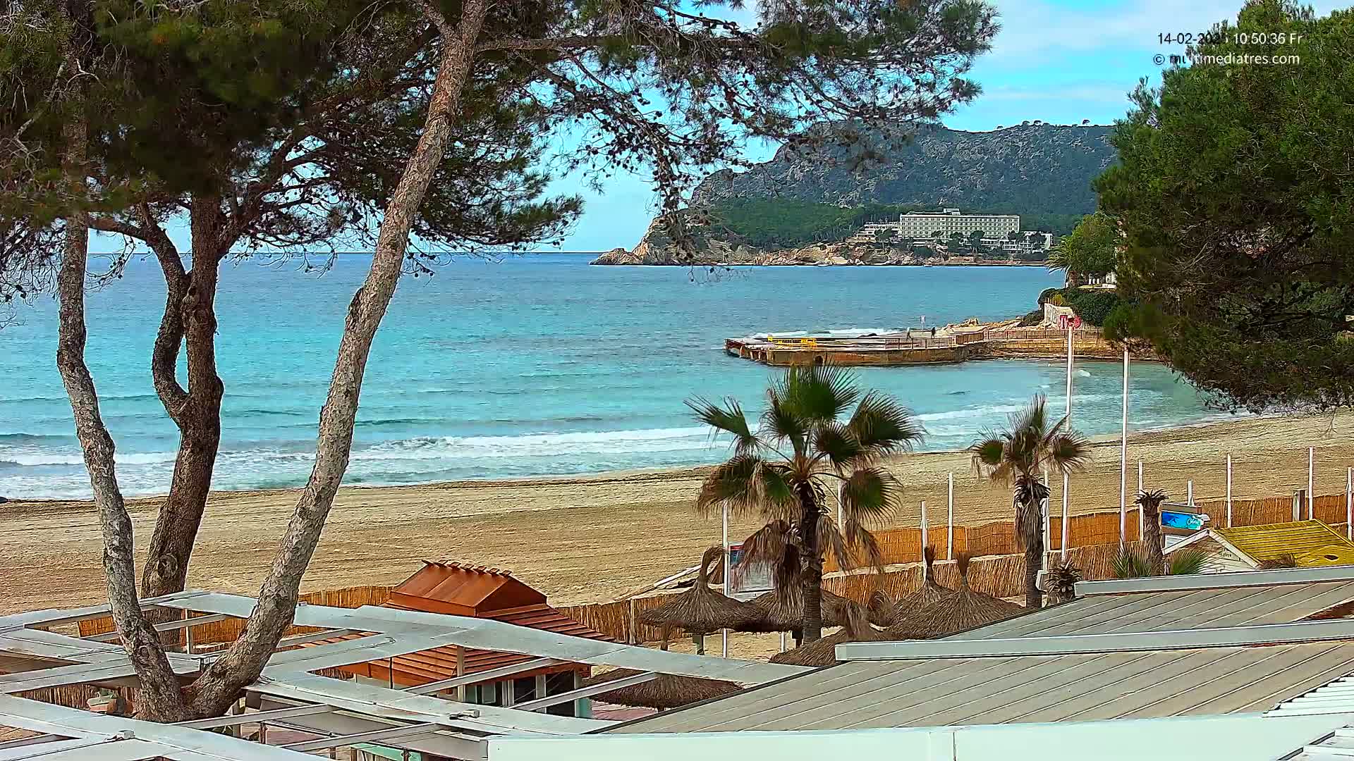 A sandy beach with calm, turquoise water is viewed from behind trees and beach structures, under a partly cloudy sky.