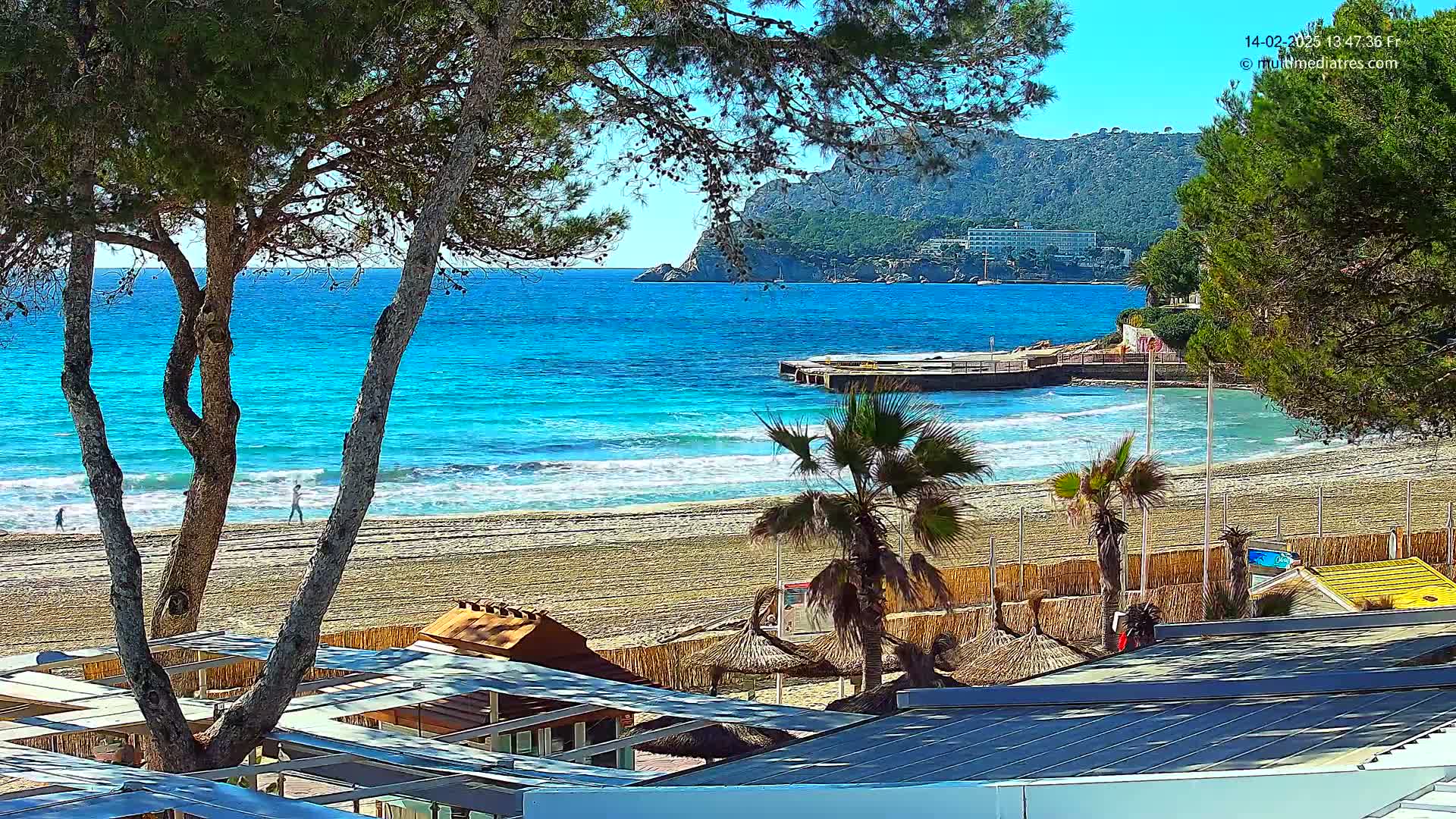 A sandy beach with turquoise water, palm trees, and beachside structures is visible under a sunny sky, with a rocky hill visible in the background.