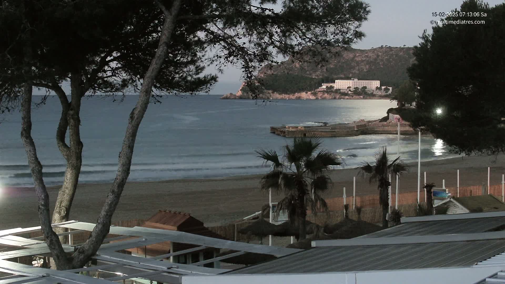 A sandy beach with calm waves and a large hotel in the background is seen at dusk, under a mostly clear sky.