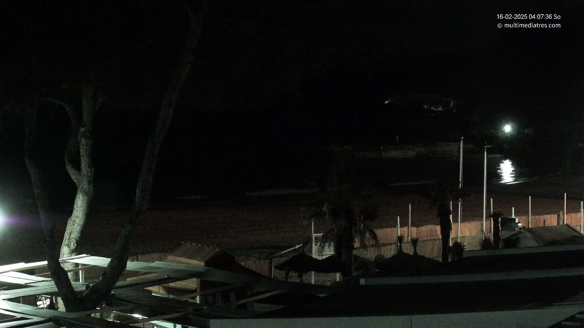 A dark, nighttime scene shows a beach with calm water reflecting light, a few palm trees, and structures near the water, all under a mostly clear night sky.