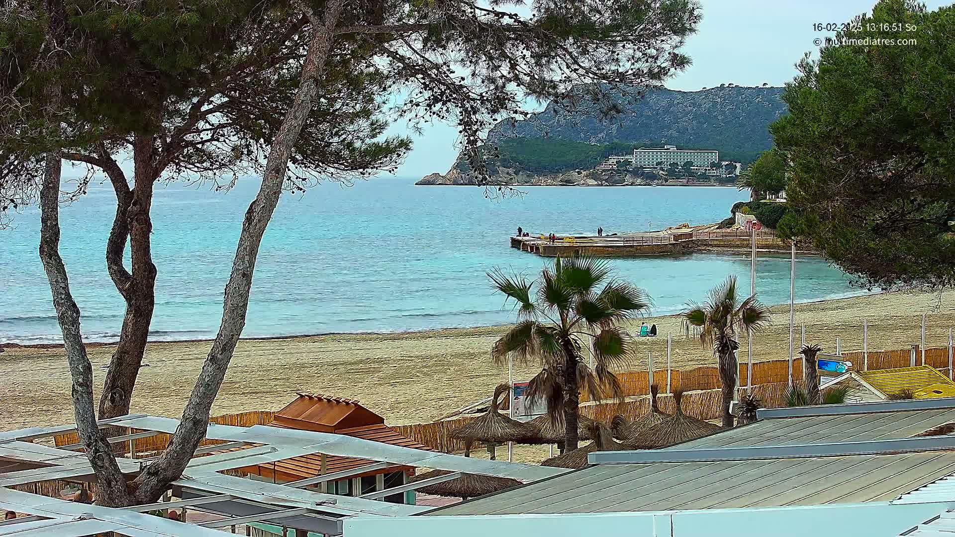 A sandy beach with turquoise water, palm trees, and beach huts is seen from above on an overcast day, with a large hotel visible on a distant hillside.