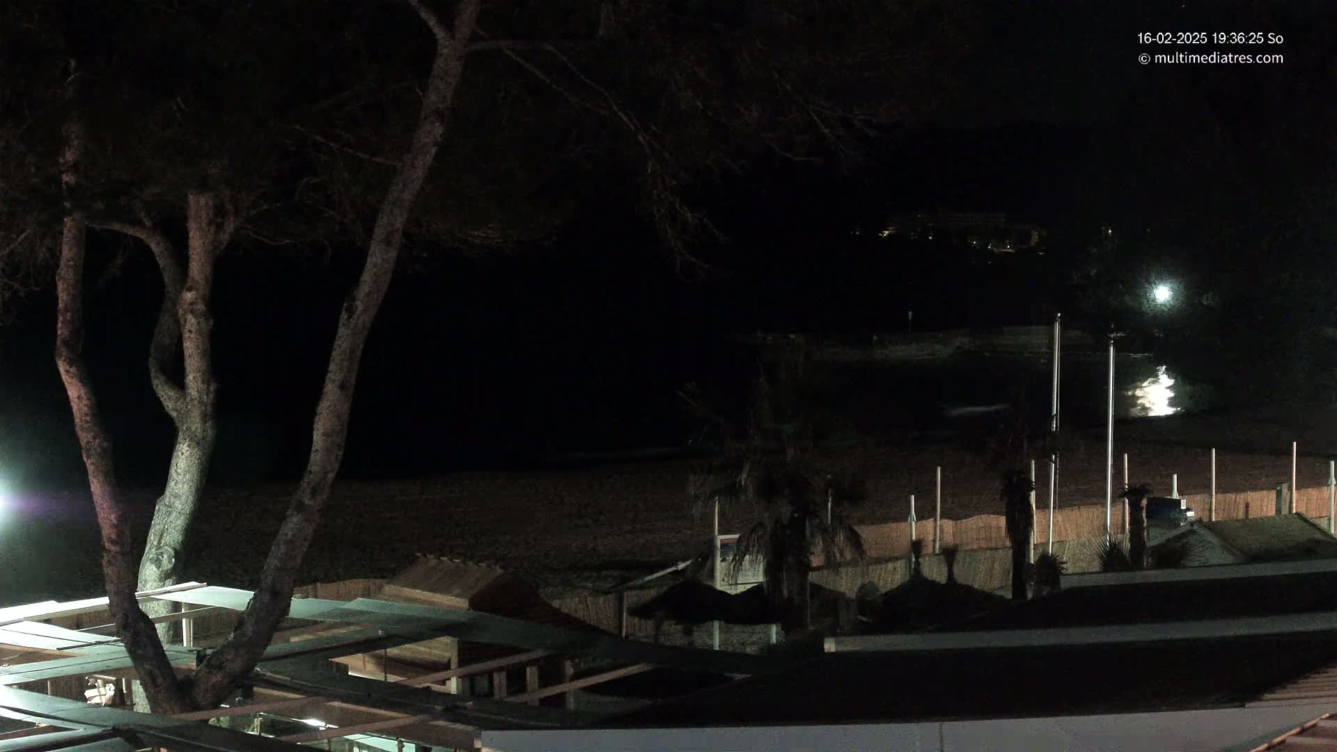 A nighttime view of a dark beach with a tree in the foreground, several structures with thatched roofs, and a dimly lit body of water in the background.