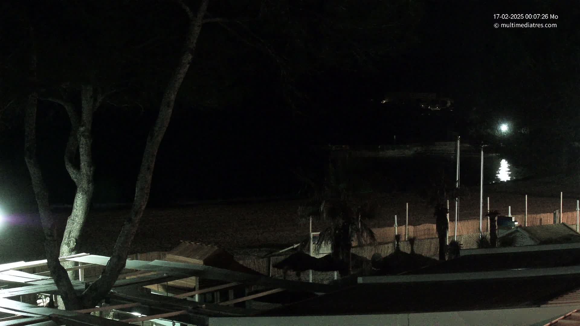 A nighttime view of a dark beach with a tree in the foreground and several beach huts and palm trees silhouetted against a calm, reflective body of water.