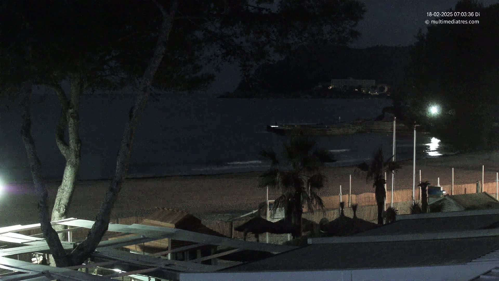 A dark nighttime beach scene shows a calm sea under a mostly clear sky, palm trees, and beach structures silhouetted against the sand.