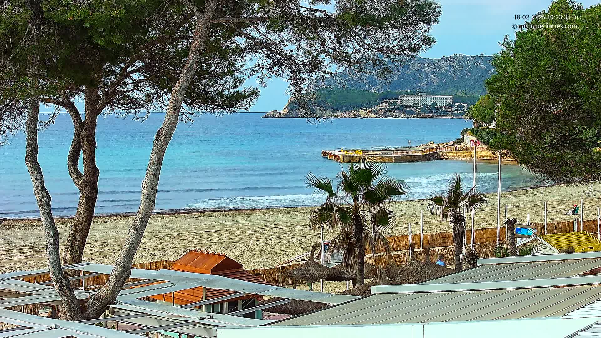 A sandy beach with calm, light blue water, palm trees, and beachside structures is seen from above on a sunny day with a hotel visible in the distance.