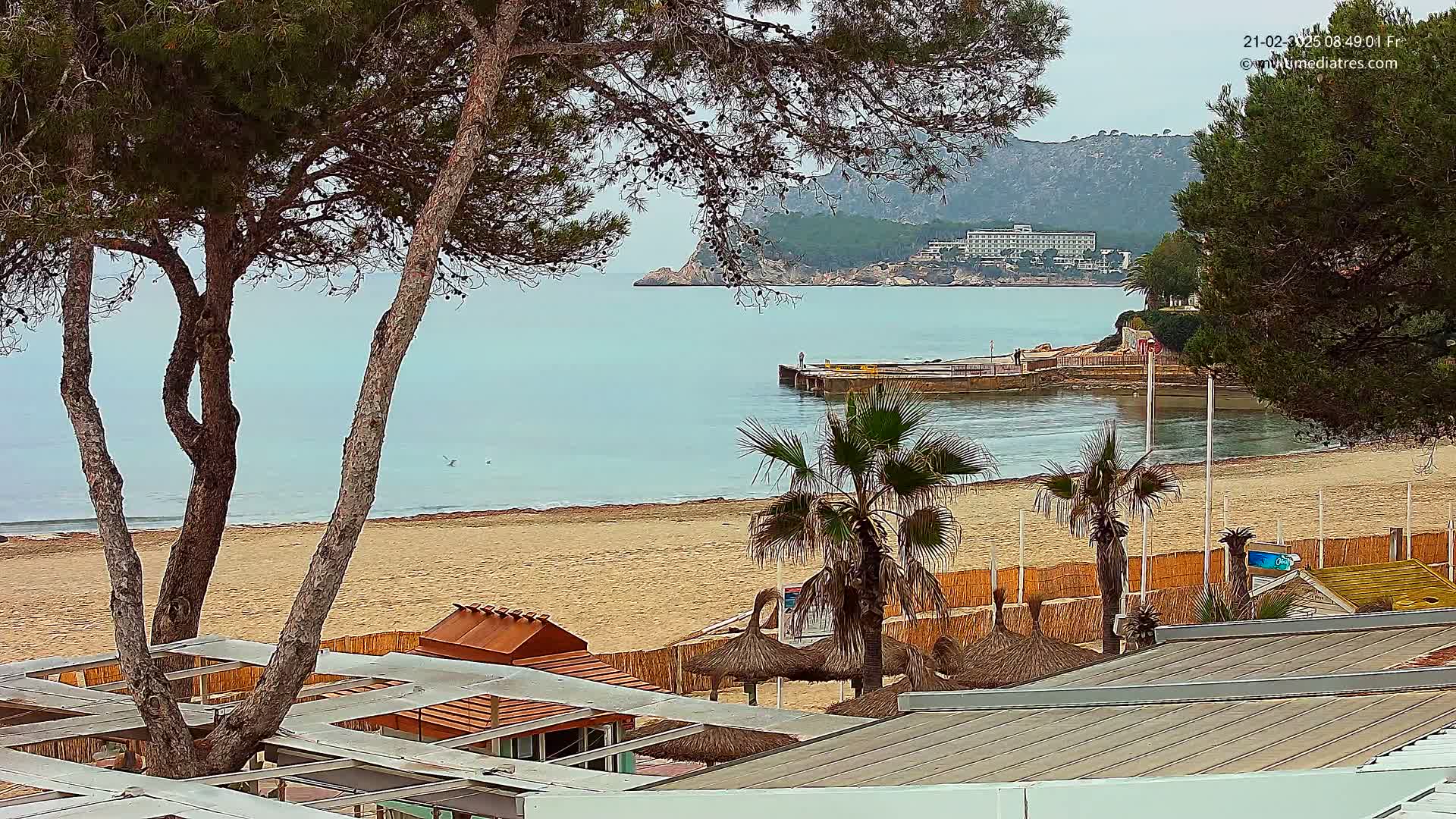 A sandy beach with calm, light-blue water, palm trees, and several small structures is seen from an elevated vantage point on an overcast day, with a distant hotel visible across the bay.