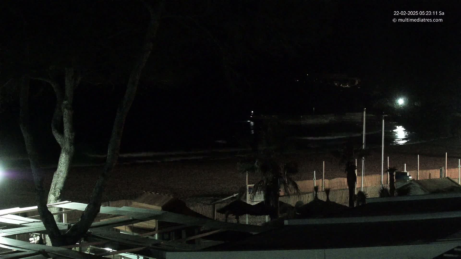 A dark nighttime beach scene shows a dark ocean with small waves lapping a sandy shore, with palm trees and beach structures visible under dim lighting.