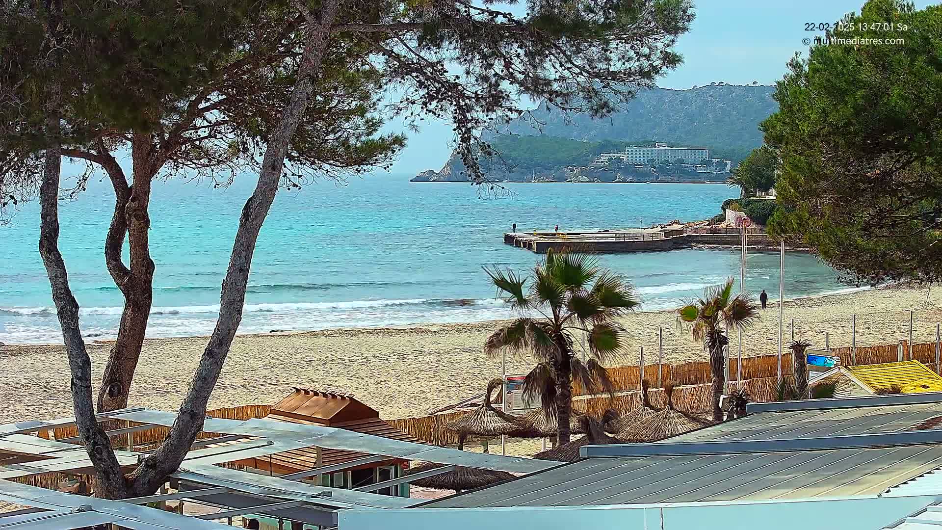 A sandy beach with turquoise water, palm trees, and beachside structures is visible under a sunny sky.