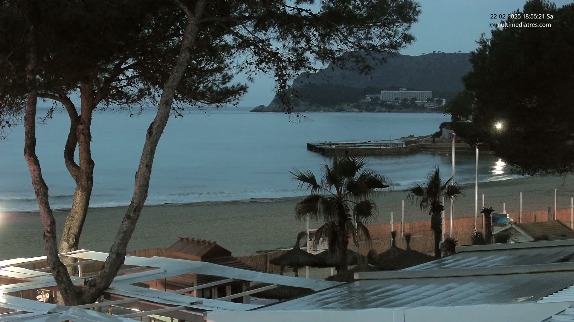 A sandy beach at dusk, with palm trees and beach structures visible, overlooks a calm sea under a dark sky.