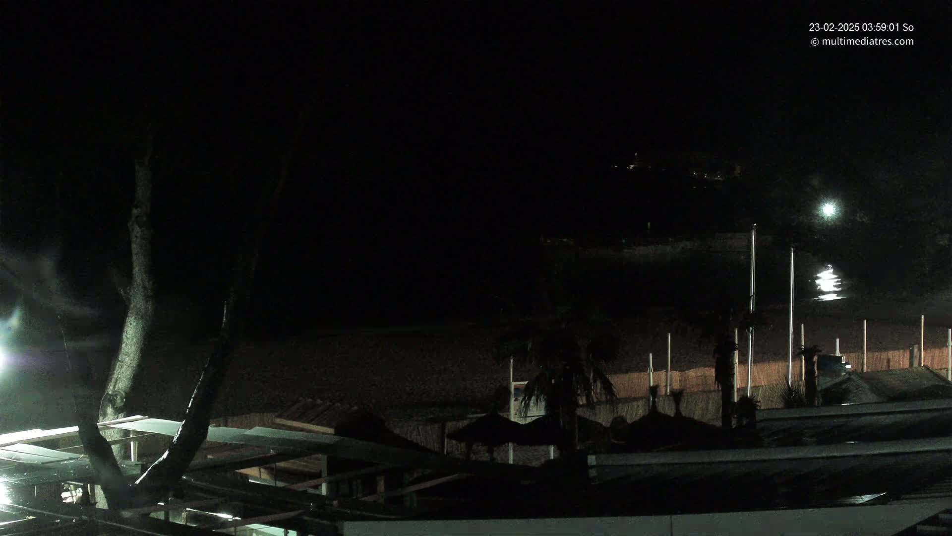 A dark, nighttime view of a beach with structures and palm trees, under a clear sky.