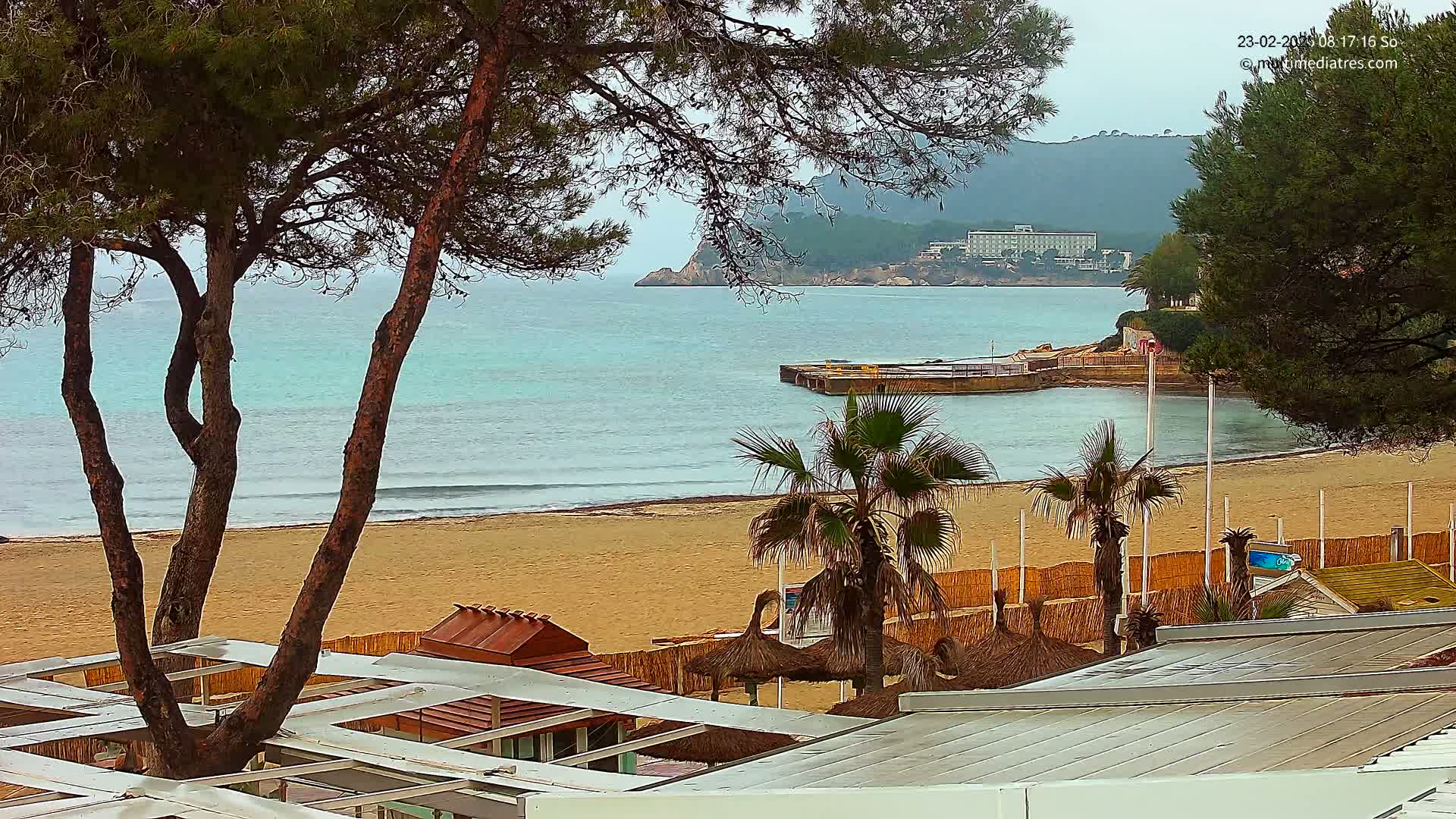 A sandy beach with calm, light-blue water, palm trees, and beach structures is seen from an elevated vantage point under an overcast sky.