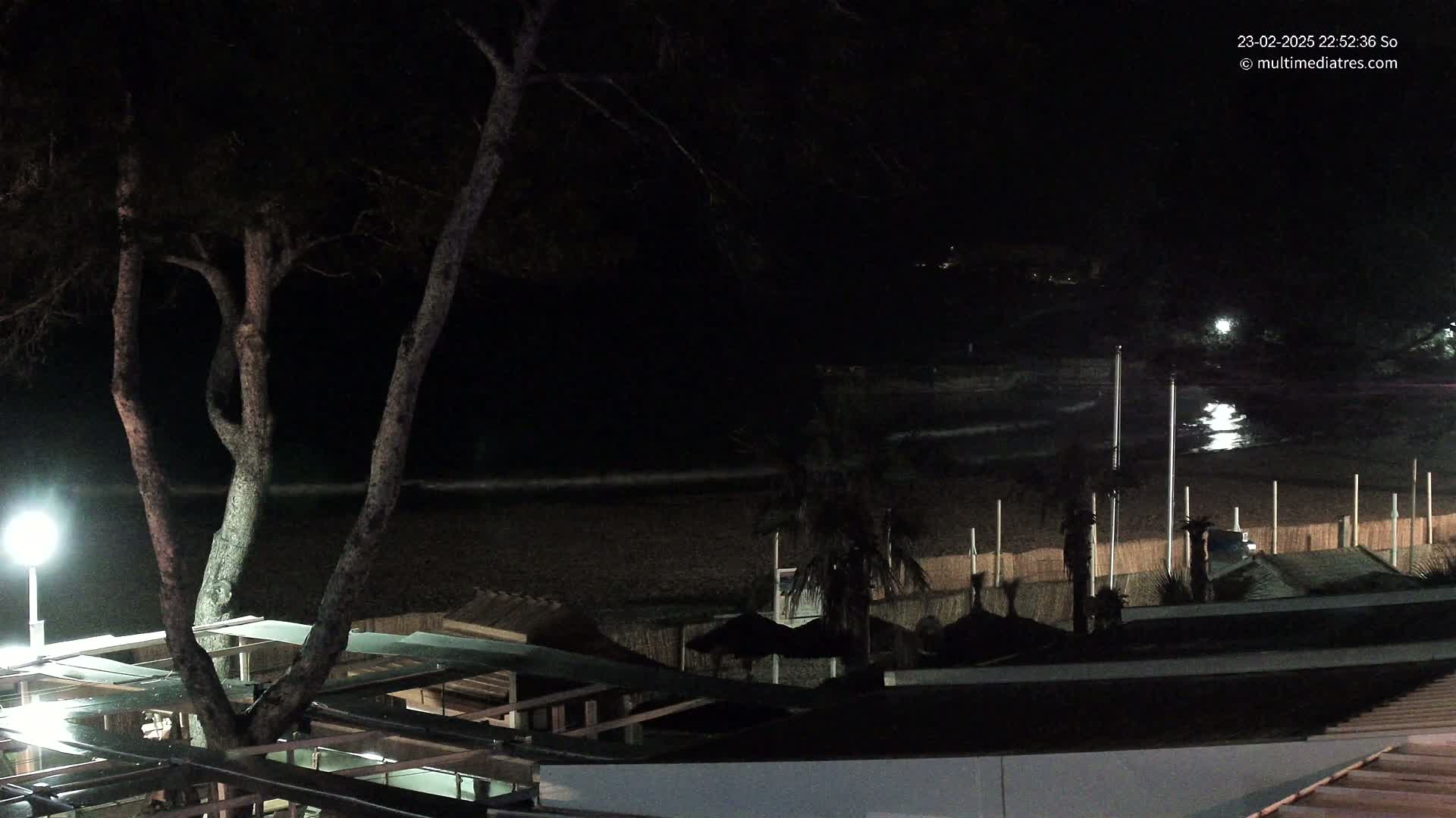 A nighttime view of a dark sandy beach with gentle waves, palm trees, and beach structures under a clear, dark sky.