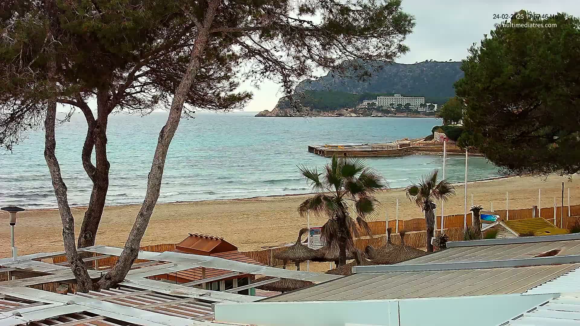 A sandy beach with calm waves laps the shore under an overcast sky, palm trees and beach structures line the sand, and a hotel is visible on a distant hillside.
