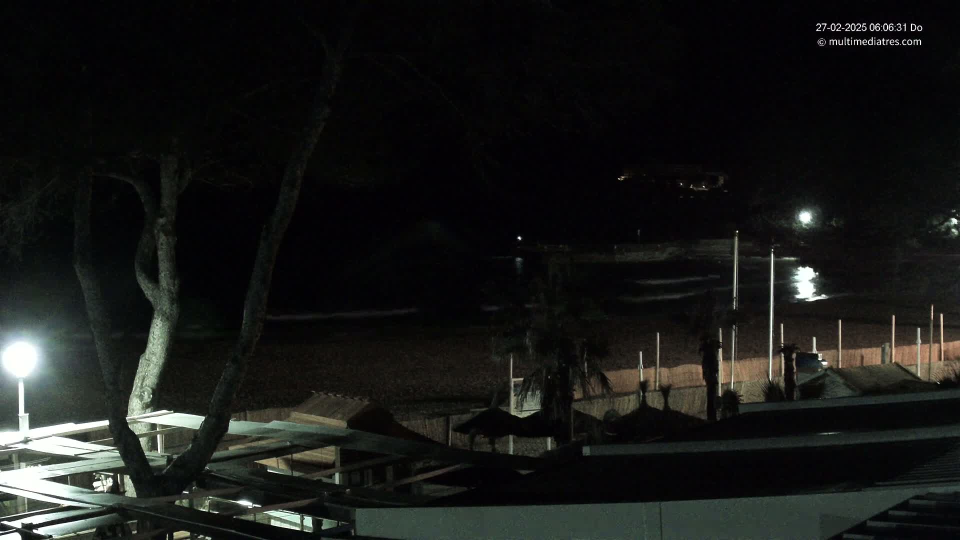 A nighttime view of a dark beach with calm waves, sparsely lit beach structures, and a tree silhouetted against the water.