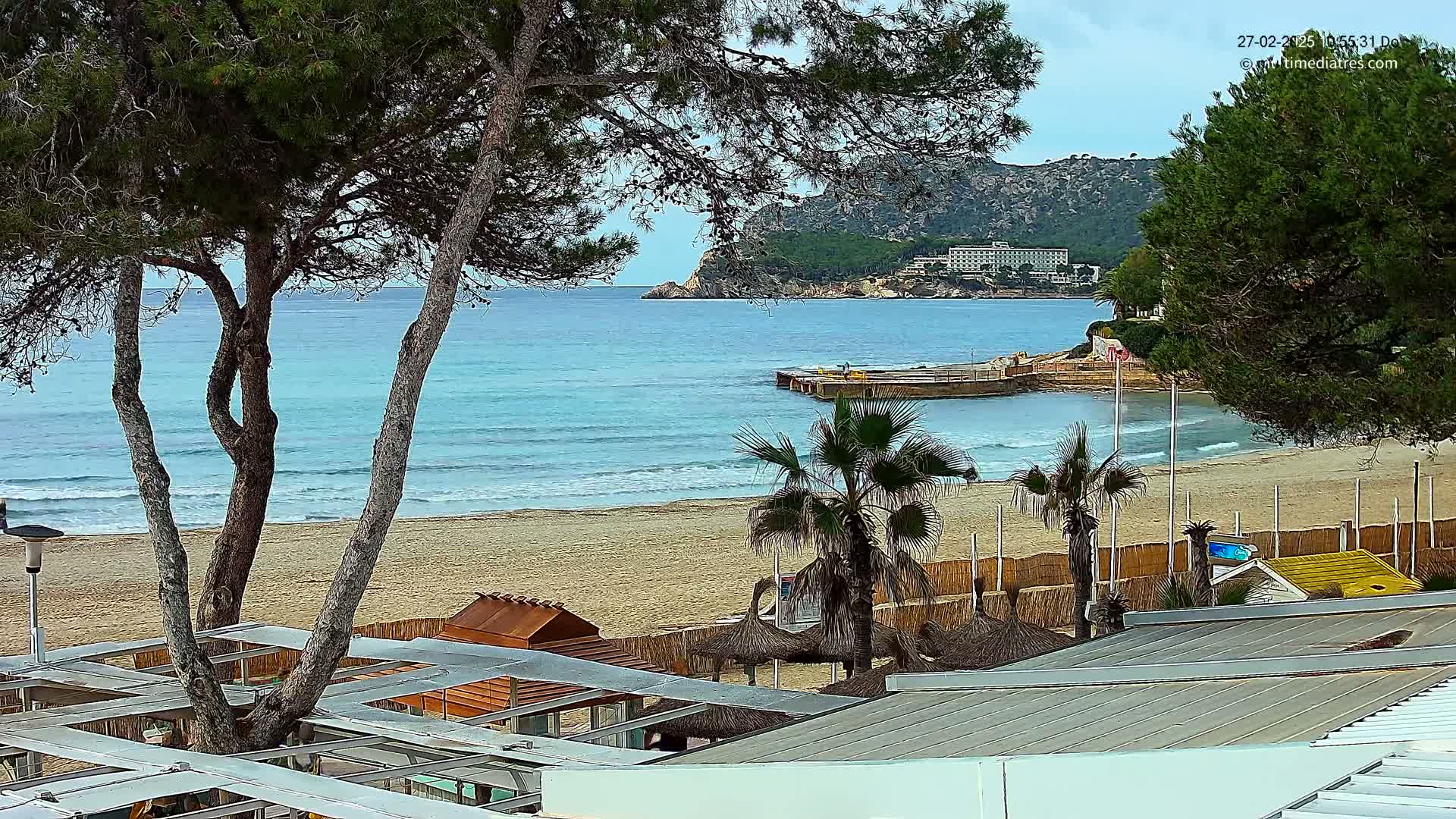 A sandy beach with calm waves and turquoise water is seen from behind a row of beach huts under a mostly cloudy sky, with trees, palm trees, and a distant hotel in view.