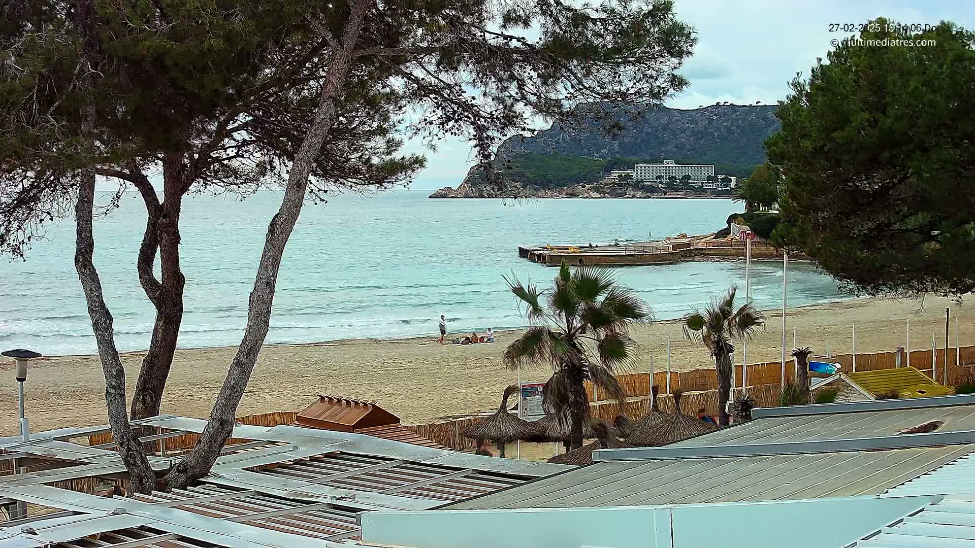 A sandy beach with calm, light blue water is partially obscured by trees, with a few people visible on the sand and a distant building on a hillside.