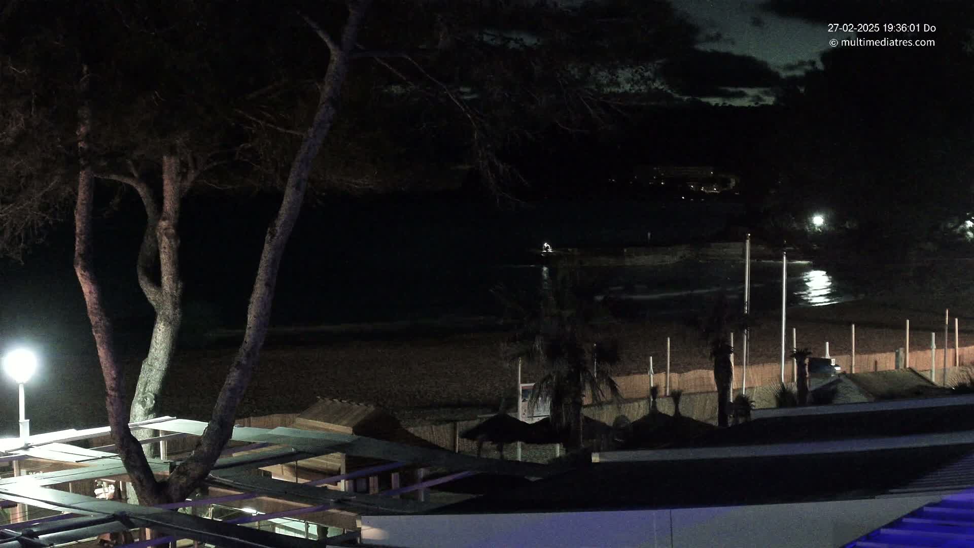 A nighttime view of a dark beach with calm water, palm trees, and beach structures under a partly cloudy sky.