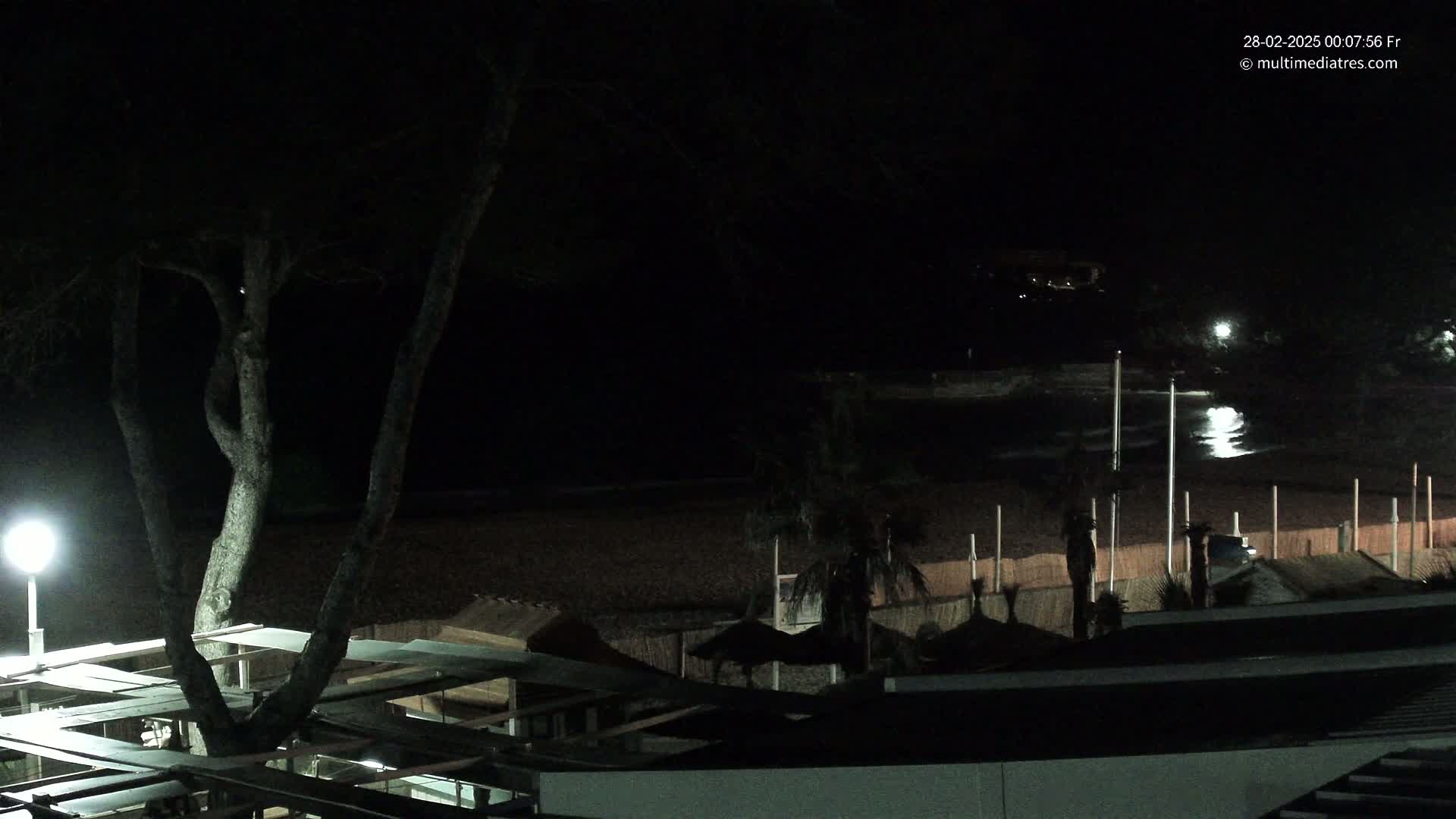 A nighttime view of a dark beach with a calm sea, partially illuminated structures, and trees under a clear sky.