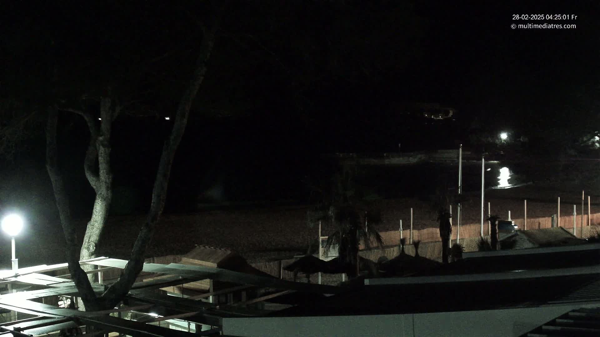 A dark, nighttime view of a beach with a few lights, palm trees, and structures visible under a clear sky.
