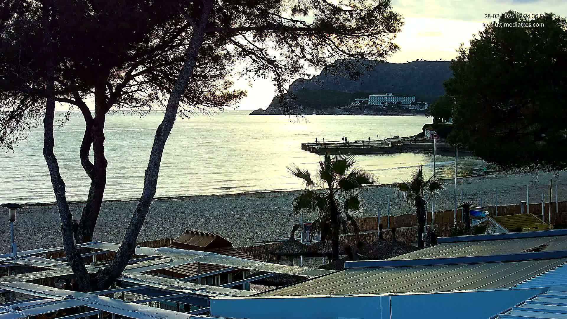 A beach scene at dusk shows a calm sea, a sandy shore, palm trees, beach structures, and a rocky cliff with buildings in the background.
