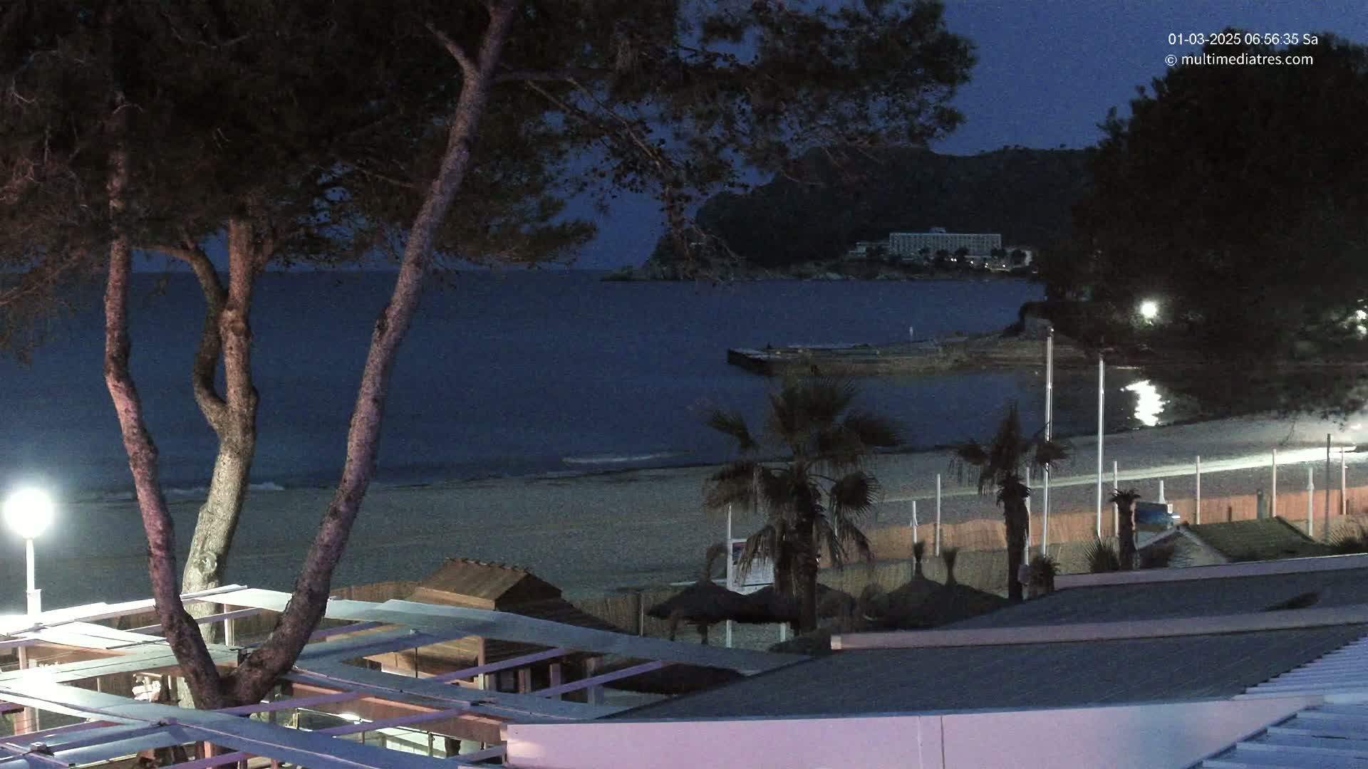 A nighttime view of a calm, dark-blue ocean beach with palm trees, beach umbrellas, and buildings under a twilight sky.