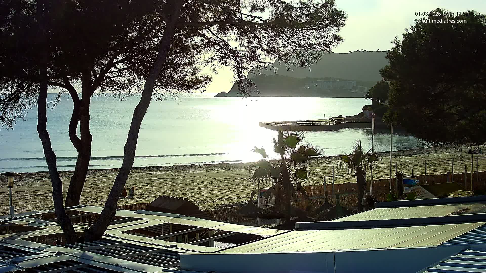 A sandy beach with calm ocean waves under a partly sunny sky is viewed from behind a structure with a slatted roof, several palm trees are visible, and a rocky hill is in the distance.