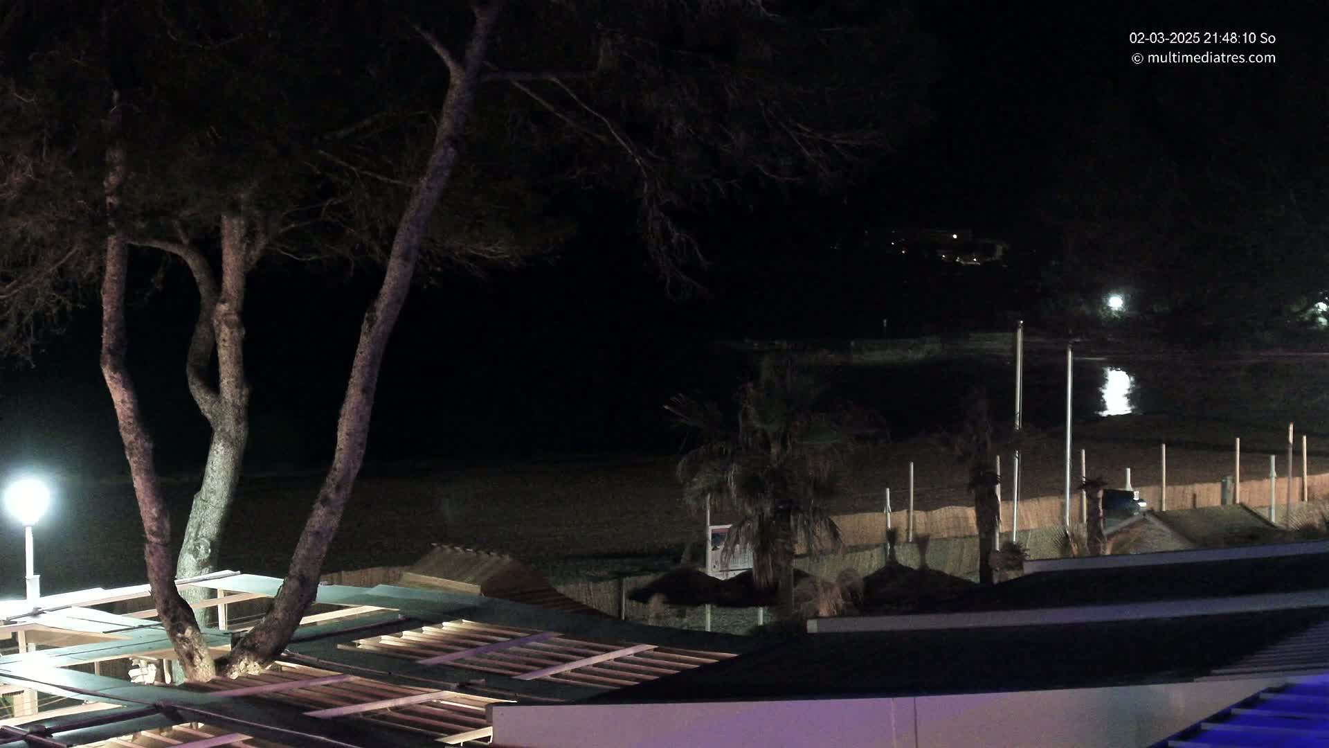 At night, a dark beach with calm water is seen from above, beyond a structure with a slatted roof and several trees.
