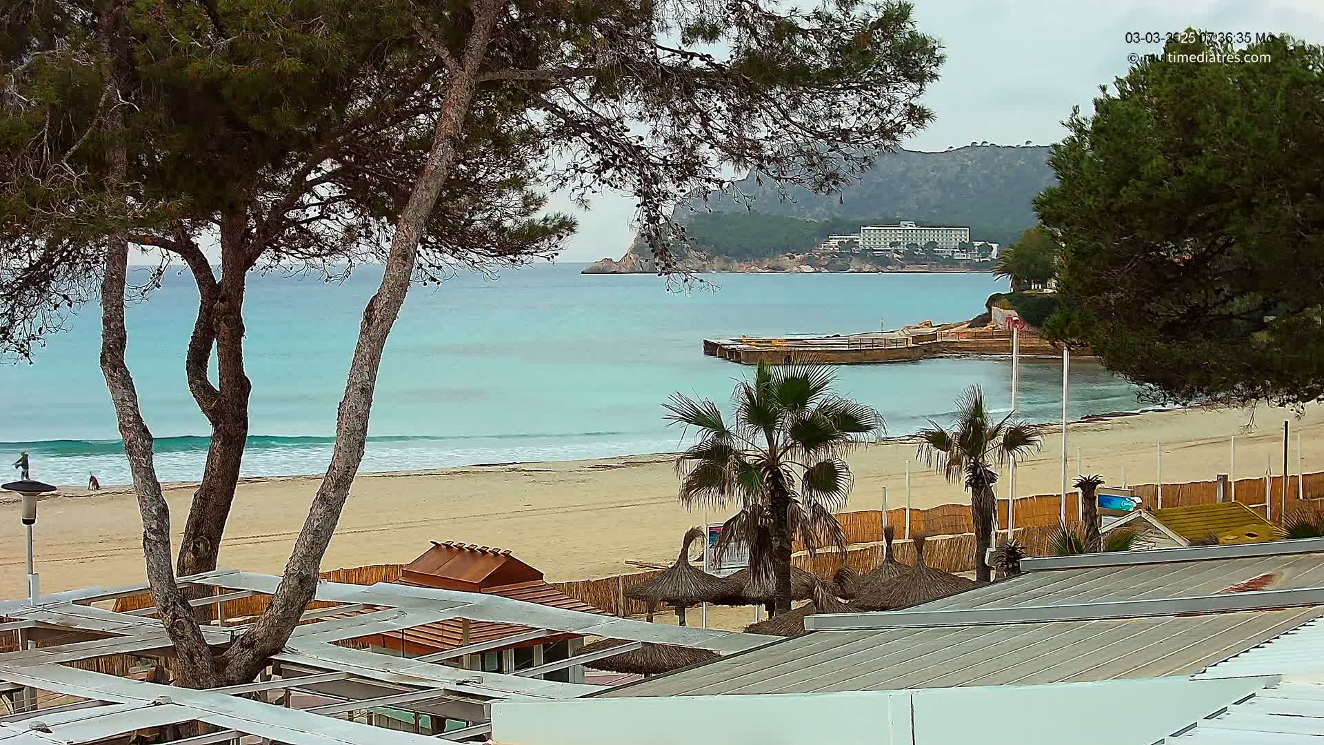 A sandy beach with calm, teal water, palm trees, beach huts, and a distant hotel, under an overcast sky.