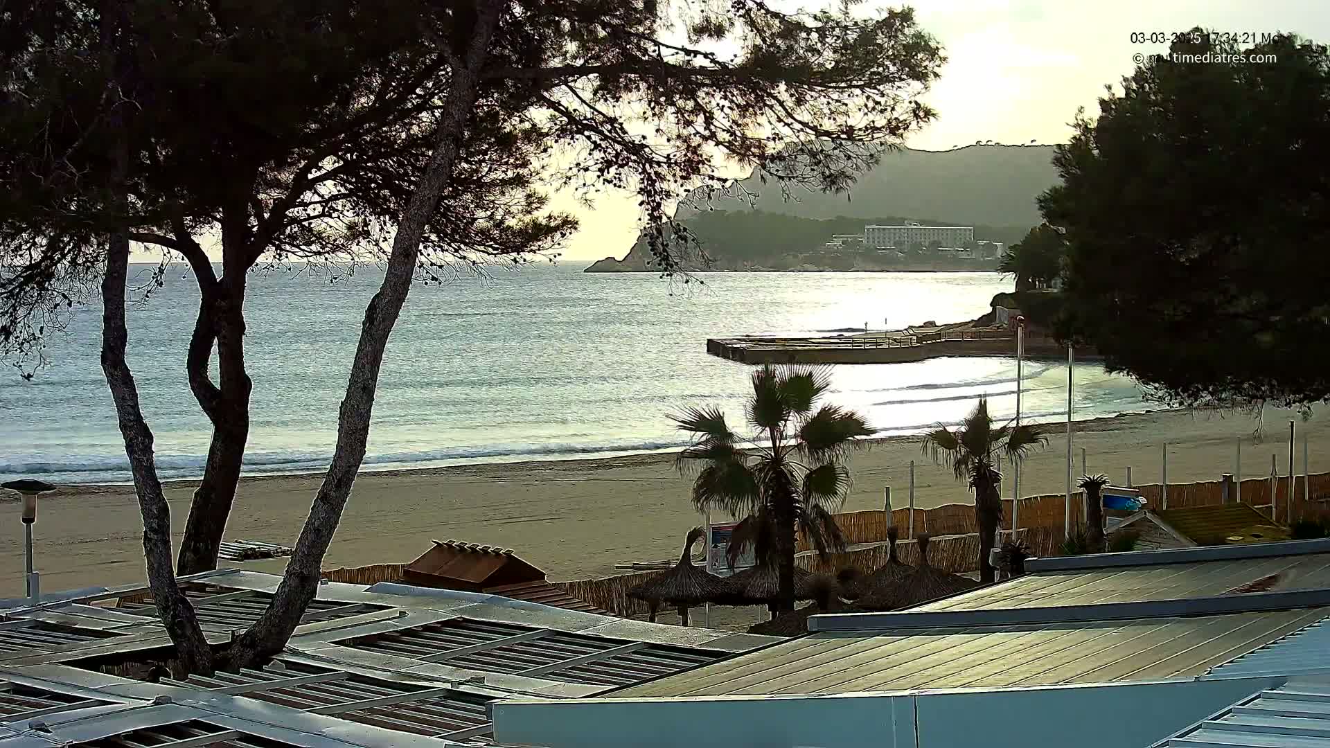 A sandy beach with calm waves and palm trees is seen from above, overlooking a calm sea and distant hills under a partly cloudy sky.