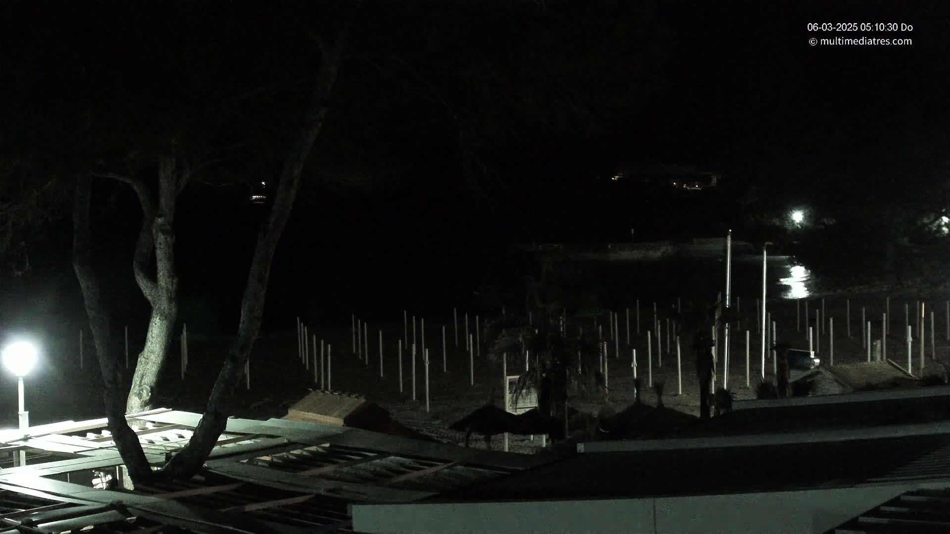 A nighttime view of a beach at night, showing a dark sandy area with numerous poles, a tree, and a building with a roof partially visible in the foreground, under a clear, dark sky.