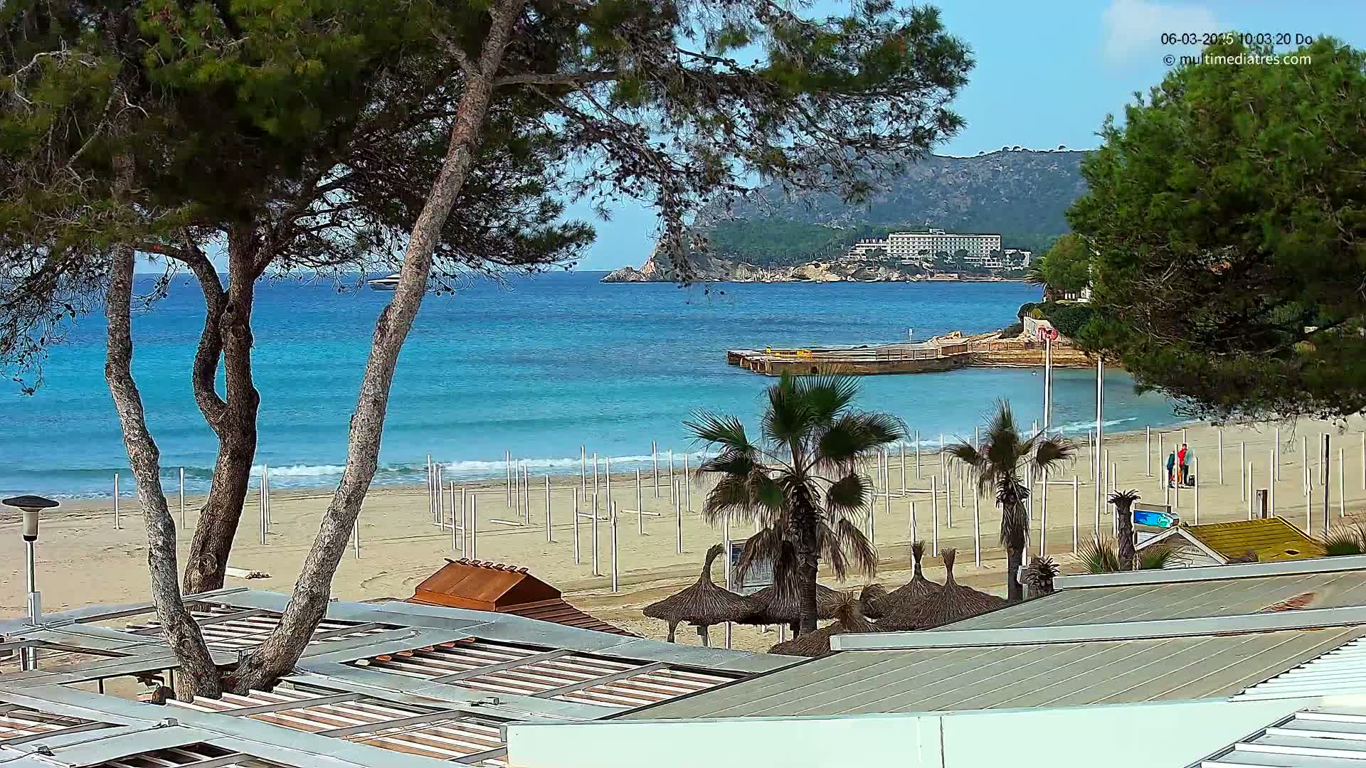 A sandy beach with turquoise water, palm trees, and a few people is seen from behind a building's roofline on a sunny day.