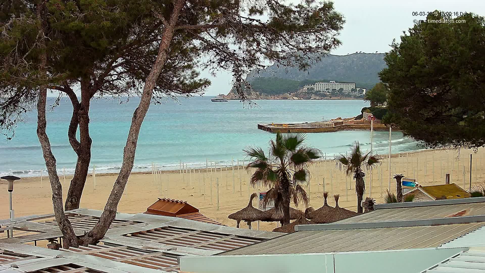 A sandy beach with turquoise water, palm trees, and beachside structures is visible under an overcast sky.