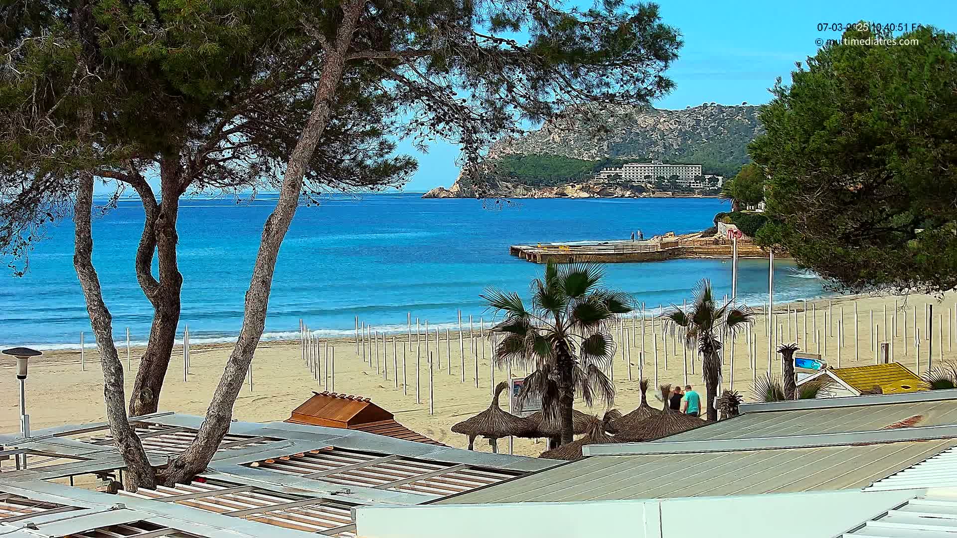 A sandy beach with turquoise water and palm trees, under a clear blue sky, is viewed from above a beachfront structure.