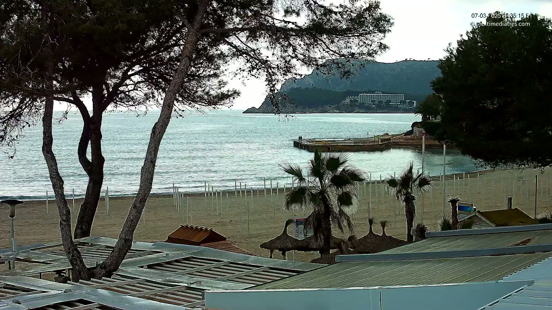 A sandy beach with palm trees and beach umbrellas is visible, overlooking a calm sea and a distant mountain under an overcast sky.