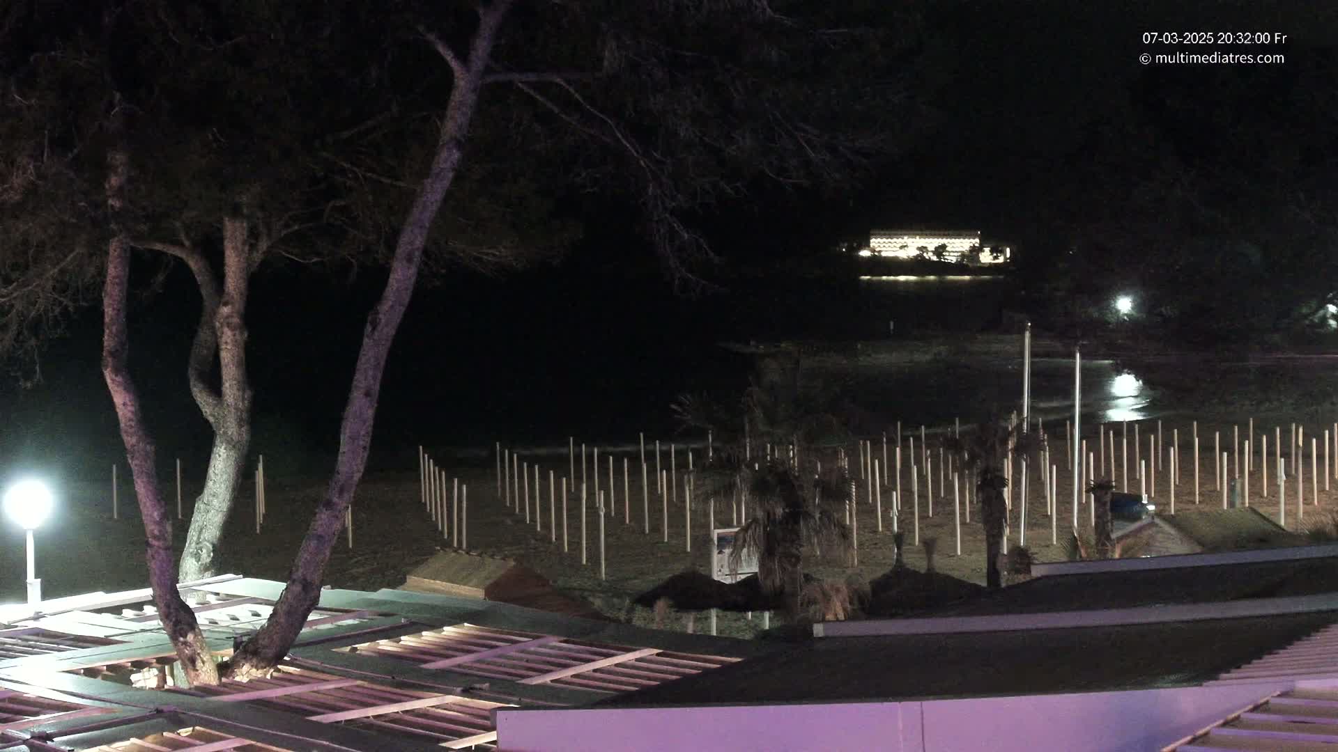 At night, a beach with many poles sticking out of the sand is visible, with a building lit up in the distance and trees in the foreground.